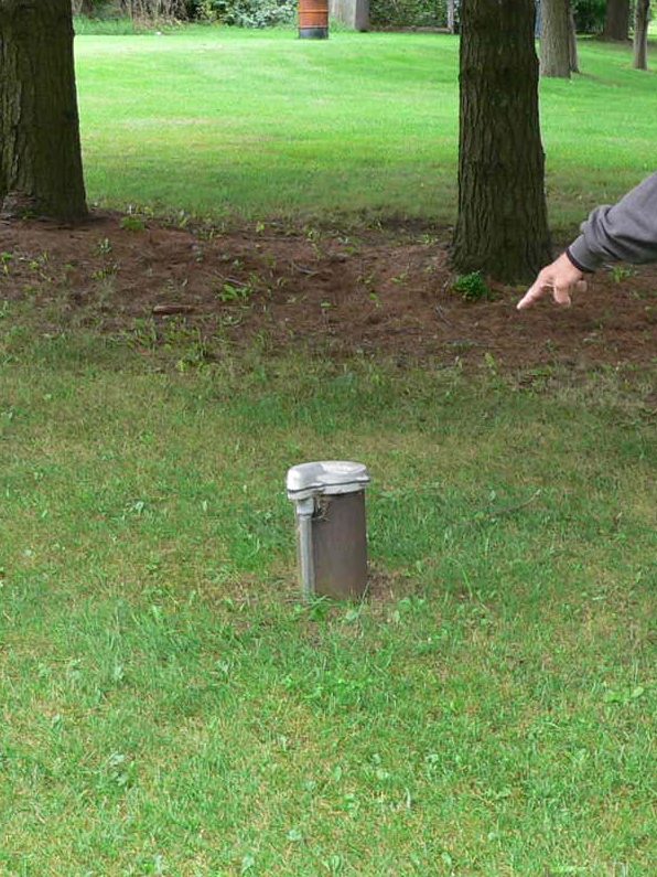 A hand points at a well cap in a grassy area.