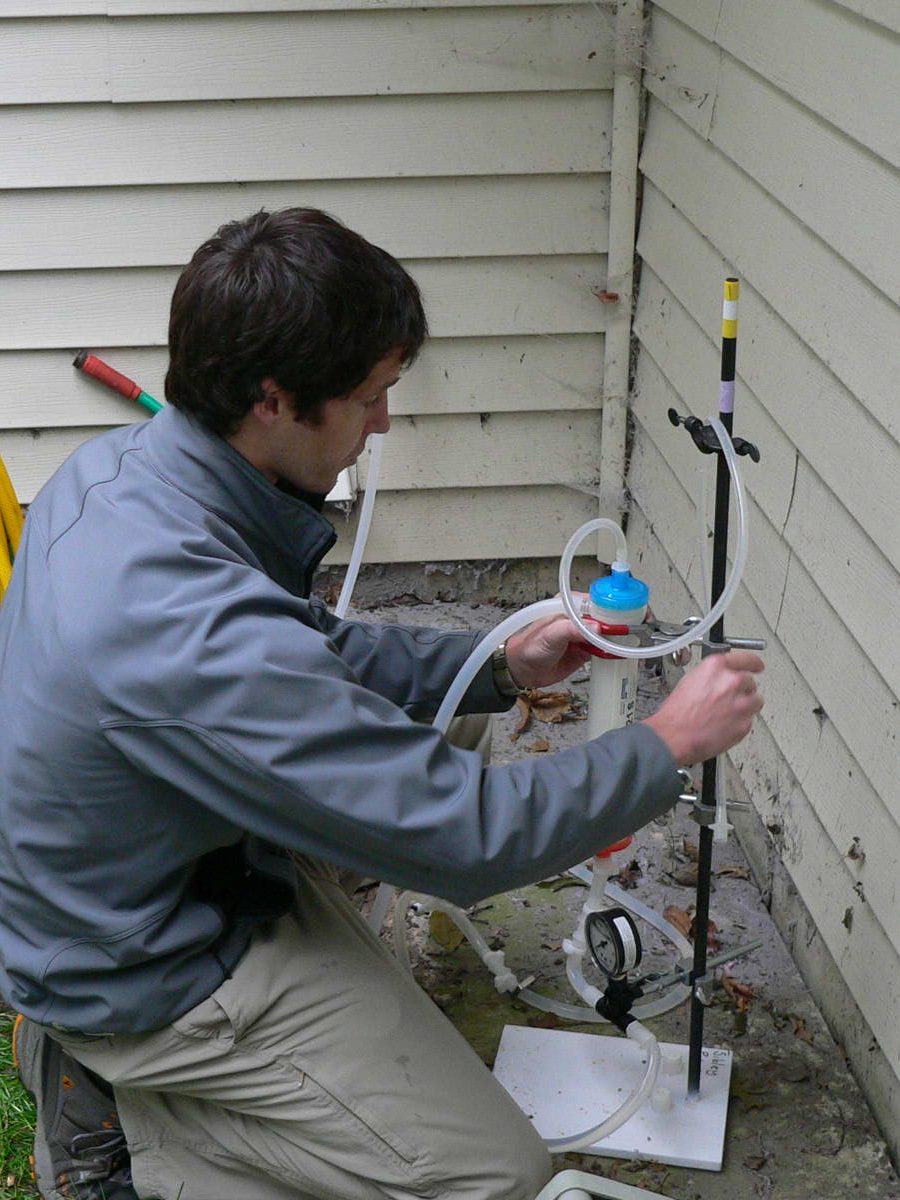 A man prepares private well testing equipment while kneeling next to the wall on the outside of the house.